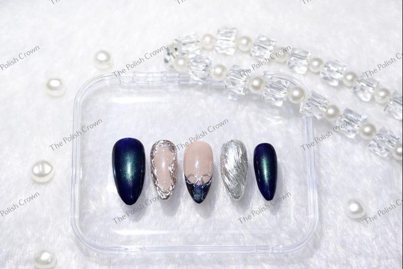 May include: A set of five artificial nails in a clear case. The nails feature various designs, including a dark blue with green shimmer, a silver textured nail, and a nude nail with silver accents. The text "The Polish Crown" is visible.