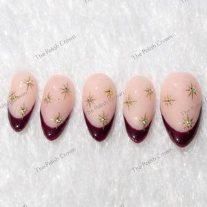 May include: Five almond-shaped artificial nails. Each nail features a light pink base with a deep burgundy tip. Gold star-shaped embellishments are scattered across the pink surface. The Polish Crown is printed on the background.