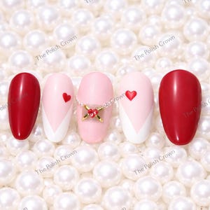 May include: An assortment of artificial nails with various designs. Two nails are solid red, while others feature pink and white patterns, some with small red heart accents. One nail has a decorative gold and red embellishment. The nails are set against a backdrop of white pearls.