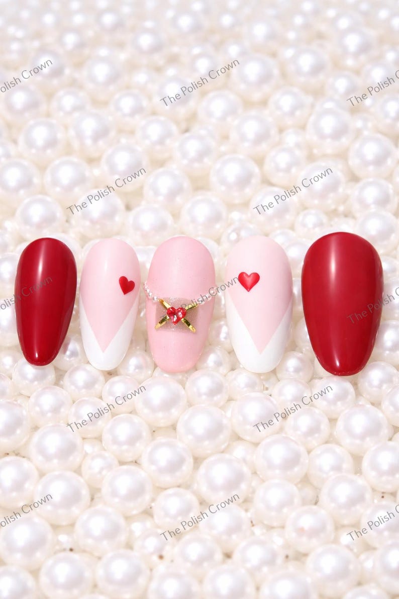 May include: An assortment of artificial nails with various designs. Two nails are solid red, while others feature pink and white patterns, some with small red heart accents. One nail has a decorative gold and red embellishment. The nails are set against a backdrop of white pearls.