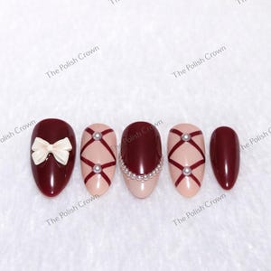 May include: A set of five artificial nails. The nails are oval-shaped and painted in various designs. One nail features a white bow, while others have a burgundy base with pink accents, pearl embellishments, and crisscross patterns. The nails are set against a white background.