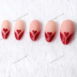 May include: Set of five artificial nails with a nude base and a red glitter heart design. The heart shape is at the tip of each nail. The nails are arranged on a white surface.
