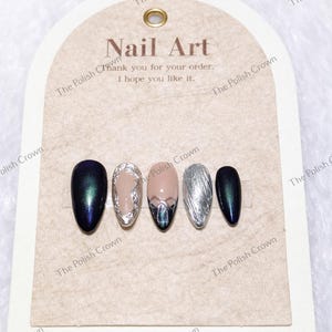 May include: A set of five artificial nails with various designs. The nails feature colors like navy, silver, and nude, with some adorned with intricate silver and iridescent accents. The card reads "Nail Art" and "Thank you for your order. I hope you like it."