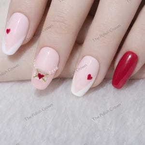 May include: A hand with manicured nails. Some nails are light pink with white tips and red hearts. One nail features a small envelope design with a heart. Another nail is solid red.