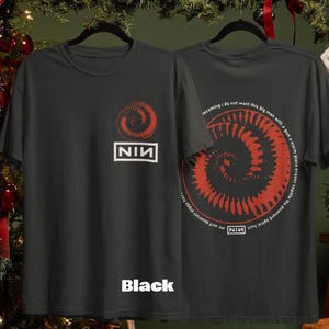 Op de afbeelding: Zwart T-shirt met een rood spiraalontwerp en de tekst "NIN" in een witte rechthoek. De achterkant van het shirt heeft een vergelijkbaar spiraalontwerp met tekst. Het woord "Black" staat onderaan het shirt.
