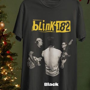 May include: Black t-shirt featuring the band Blink-182's logo in yellow, with a black and white image of the band members. The word "Black" is printed at the bottom of the shirt.