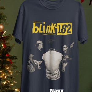 May include: Navy blue t-shirt featuring the band Blink-182's logo in yellow. The shirt has a graphic of the band members playing instruments. The word "Navy" is printed at the bottom of the shirt.