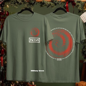 May include: Military green t-shirts with a red spiral design and the text "NIN". One shirt displays the NIN logo and the text "Military Green". The other shirt has a spiral design with text around it.