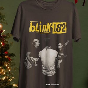 May include: Dark chocolate t-shirt featuring the band Blink-182. The shirt has a yellow rectangular logo with the band's name and number. A black and white image of the band members playing their instruments is on the front.