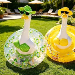 May include: Two inflatable swan pool floats on a grassy lawn. One swan wears a green polka-dot hat, sunglasses, and bikini. The other swan wears a yellow hat, sunglasses, and bikini. Both swans have white necks and yellow beaks.