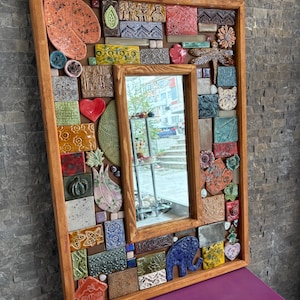 Könnte beinhalten: Ein rechteckiger Spiegel mit Holzrahmen, einem zentralen Spiegel und einem Mosaikrand. Das Mosaik zeigt verschiedene farbenfrohe Keramikfliesen mit floralen, tierischen und geometrischen Designs. Der Rahmen hat eine warme braune Farbe.