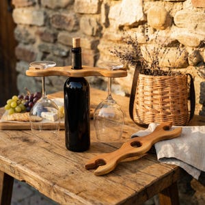 以下が含まれることがあります： 2つのグラスホルダー、ワインボトル、ワイングラス2つが付いた木製のワインボトルホルダー。木製のワインオープナーがテーブルにあります。ラベンダーが入った柳のバスケットが背景にあります。テーブルは木製です。