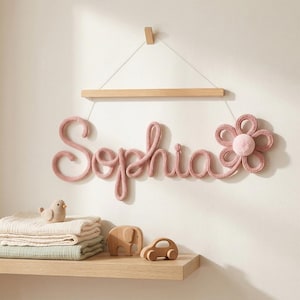 K&ouml;nnte beinhalten: Rosa gestrickte Wanddekoration mit dem Namen "Sophia" und einem Blumendesign. Der Name h&auml;ngt an einer Holzstange. Ein Regal darunter enth&auml;lt gefaltete Decken, einen Vogel, einen Elefanten und ein Auto aus Holz.