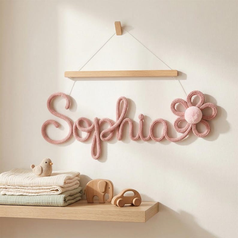 K&ouml;nnte beinhalten: Rosa gestrickte Wanddekoration mit dem Namen "Sophia" und einem Blumendesign. Der Name h&auml;ngt an einer Holzstange. Ein Regal darunter enth&auml;lt gefaltete Decken, einen Vogel, einen Elefanten und ein Auto aus Holz.