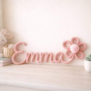 K&ouml;nnte beinhalten: Rosa gestrickte Wanddekoration mit dem Namen "Emma" in Schreibschrift, begleitet von einer Blume mit einem zentralen Pompon. Die Dekoration befindet sich auf einer hellen Oberfl&auml;che vor einer wei&szlig;en Wand und erzeugt eine weiche, einladende &Auml;sthetik.