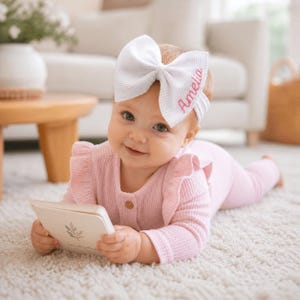 May include: A white waffle-textured fabric hair bow featuring the embroidered name Amelia in pink script, attached to a white elastic headband. The bow is worn on the head, paired with a light pink long-sleeved ribbed romper with ruffled shoulders.