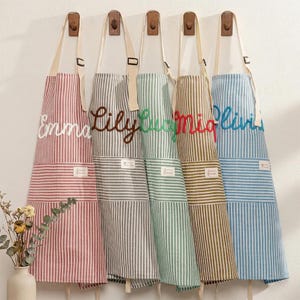 Puede incluir: Cinco delantales a rayas en rojo, gris, verde, marrón y azul cuelgan de ganchos de madera. Cada delantal tiene un nombre bordado diferente: Emma, Lily, Lucy, Mia y Olivia. Los delantales tienen una correa blanca y un bolsillo.