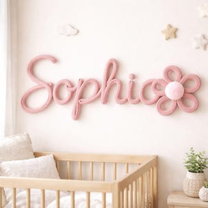 K&ouml;nnte beinhalten: Rosa gestrickte Wanddekoration mit dem Namen "Sophia" in Schreibschrift, begleitet von einer passenden Blume. Die Kinderzimmerdekoration umfasst ein Holzbett, Kissen und sternf&ouml;rmige Wandakzente, die eine weiche, einladende Atmosph&auml;re schaffen.