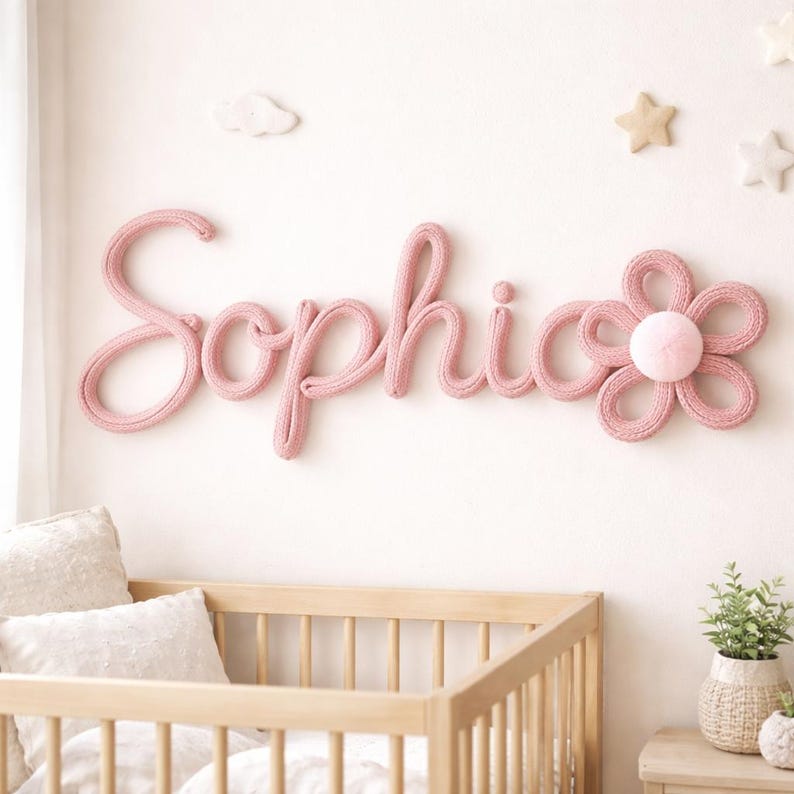 K&ouml;nnte beinhalten: Rosa gestrickte Wanddekoration mit dem Namen "Sophia" in Schreibschrift, begleitet von einer passenden Blume. Die Kinderzimmerdekoration umfasst ein Holzbett, Kissen und sternf&ouml;rmige Wandakzente, die eine weiche, einladende Atmosph&auml;re schaffen.