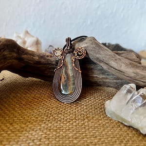May include: A handcrafted copper wire pendant with a teardrop-shaped stone. The stone features shades of blue, brown, and tan. The pendant has intricate wire wrapping and decorative swirls at the top, suspended from a black cord.