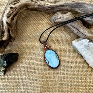 May include: A handcrafted pendant necklace featuring a light blue oval gemstone. The stone is encased in a copper wire frame and suspended from a black cord. The pendant has a decorative copper wire design at the top.