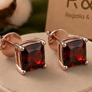 Può includere: Un paio di orecchini a lobo in oro rosa con pietre preziose granato rosso scuro a taglio quadrato. Gli orecchini sono esposti su un portagioie rotondo in velluto beige accanto a un cartoncino beige con il testo R&R Regalia & Roses e una piccola icona a piramide.