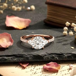 Puede incluir: Un anillo de compromiso de oro rosa con una piedra central grande, redonda y transparente y piedras de acento más pequeñas. El anillo se encuentra sobre un fondo oscuro con pétalos de rosa secos, perlas y una brújula vintage.
