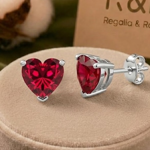 Puede incluir: Un par de pendientes de botón de plata con piedras rojas facetadas en forma de corazón. Los pendientes se muestran sobre un soporte redondo de color beige junto a una tarjeta con el texto R&R Regalia & Roses.