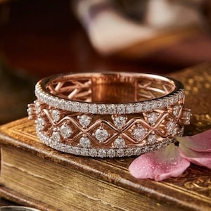 Könnte beinhalten: Roségoldener Ring mit Diamantbesatz. Der Ring hat eine Reihe von Diamanten in einem sich wiederholenden Muster, wodurch ein funkelnder Effekt entsteht. Der Ring liegt auf einem alten Buch mit einer Lupe.