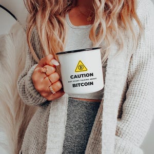 Puede incluir: Taza de esmalte blanco con borde negro con el texto "CAUTION MAY START TALKING ABOUT BITCOIN" y un triángulo amarillo con un símbolo de Bitcoin. La persona lleva un cárdigan gris.