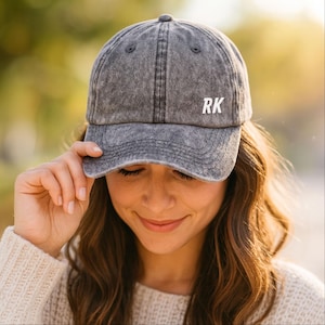 May include: A gray, distressed baseball cap with the white letters "RK" on the front. The cap is worn by a person with long brown hair and a cream-colored sweater. The background is blurred with natural light.