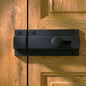 May include: A black door bolt latch with a circular handle and a sliding mechanism. The latch is mounted on a light brown wooden door with visible wood grain and is secured with screws.