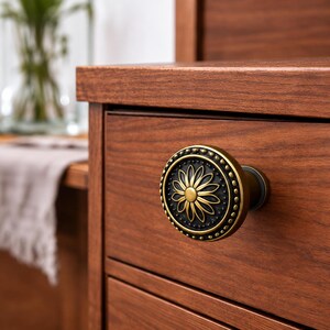 Peut inclure: Un bouton de meuble rond présentant un centre noir foncé avec un motif de pétales de fleur doré en relief. Le bord extérieur du bouton est orné d'une bordure perlée décorative dans un ton doré assorti, fixé sur une façade de tiroir en bois.