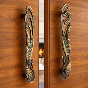 May include: A pair of vertical door handles crafted from antique brass-toned metal, each featuring a stylized peacock design with an elongated tail. The handles are embellished with small, inset emerald green stones along the curved metal plumes.