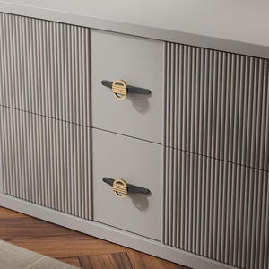 May include: A light grey cabinet featuring two central drawers with smooth faces and two side sections with vertical fluted paneling. Each drawer is equipped with a horizontal handle consisting of a black bar and a circular gold-toned accent.