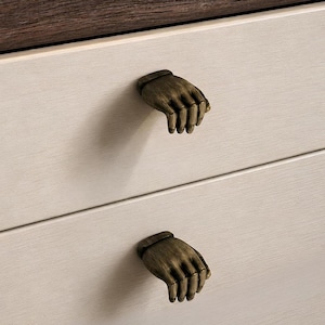 May include: Two drawer pulls shaped like human hands in an antique brass finish. The hand-shaped knobs are mounted on a light cream-colored drawer front with a subtle horizontal wood grain texture, positioned below a dark brown wooden surface.