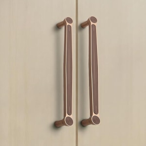 Könnte beinhalten: Zwei vertikale Schrankgriffe mit metallischem Kupfer-Finish. Jeder Griff verfügt über eine lange, schlanke Mittelstange mit einer dunkelbraunen, gerillten, holzähnlichen Einlage. Die oberen und unteren Enden sind abgerundet und mit konzentrischen Kreisen verziert.