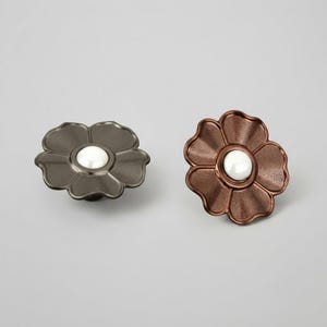 May include: Two flower-shaped cabinet knobs. One knob is dark gray with a smooth finish, and the other is a textured copper-brown. Both feature a round, white, pearl-like center accent. The knobs are circular with five rounded petals.