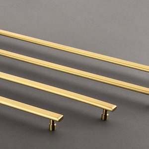 Puede incluir: Cuatro tiradores de armario de metal dorado de diferentes longitudes dispuestos en diagonal. Cada tirador presenta una barra rectangular larga y delgada con bordes biselados y dos postes de montaje cilíndricos en los extremos para su fijación.