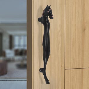 May include: A matte black metal door handle shaped like a stylized horse, featuring a detailed equine head at the top and a slender, elongated body that extends downward to a mounting point, attached vertically to a light-colored wooden door panel.
