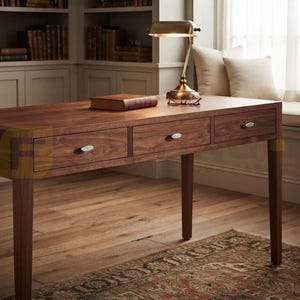 May include: A rectangular wooden desk featuring three horizontal drawers with silver-toned oval pulls. A single brown leather-bound book rests on the flat surface, accompanied by a brass-finished desk lamp with a curved arm and a wide shade.