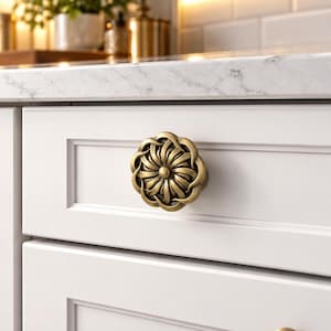 Peut inclure: Un bouton de meuble rond en laiton antique présentant un motif floral détaillé à plusieurs couches avec un bouton circulaire central. La quincaillerie est fixée sur une façade de tiroir blanche à panneau plat avec une bordure rectangulaire.