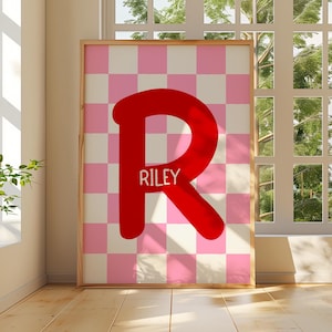 May include: A framed print with a large red letter "R" and the name "Riley" in white text. The background is a pink and white chequered pattern. The print is displayed in a room with a window.