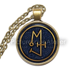 Puede incluir: Un collar de oro con un colgante redondo de vidrio azul que presenta un sello dorado. El sello es una letra estilizada "M" con un círculo y una cruz en su interior.