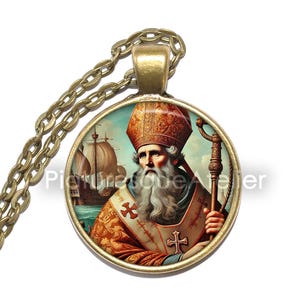 Può includere: Una collana con pendente rotondo con un'immagine dettagliata di una figura religiosa con cappello e abiti rossi e dorati, che tiene un bastone. Il pendente è su una catena dorata. Una nave in mare è sullo sfondo.