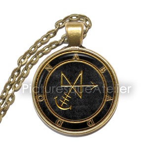 Op de afbeelding: Een gouden ketting met een zwarte en gouden hanger met een zegel met een symbool en een anker. De hanger is rond en heeft een gouden rand met de letters N, O, A, B, D en D rond de rand.