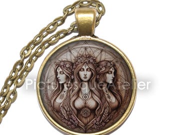 May include: A gold chain necklace with a round pendant featuring a detailed illustration of three women with floral crowns and a geometric pattern in the background.