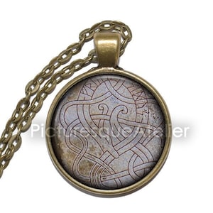 May include: A bronze chain necklace with a round pendant featuring a detailed Celtic knot design in red and white on a gray background.