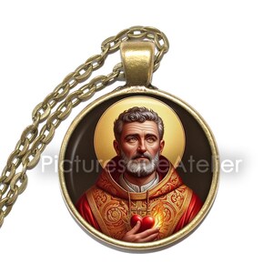 May include: A gold-coloured pendant necklace featuring a detailed image of a saint holding a heart. The pendant has a gold frame and chain. The saint is depicted with a halo, wearing a red and gold robe. The text "Picturesque Atelier" is visible.