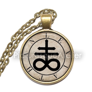 May include: A gold-toned pendant necklace featuring a circular charm with a black symbol and text. The symbol is a cross above an infinity symbol. The text around the edge reads "N L E V A T H". The chain is also gold-toned.